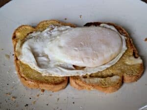 A fried egg with a slightly runny white is served on two slices of toasted bread, placed on a white plate with crumbs scattered around.