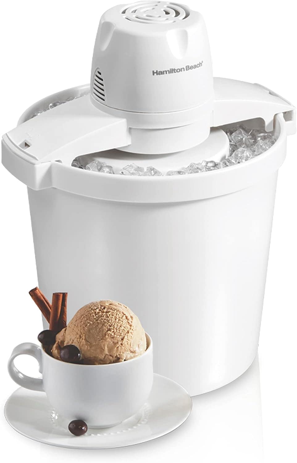 A Hamilton Beach ice cream maker filled with ice is shown with a scoop of ice cream in a white cup on a saucer in front. The ice cream cup is garnished with chocolate-covered coffee beans and two cinnamon sticks.