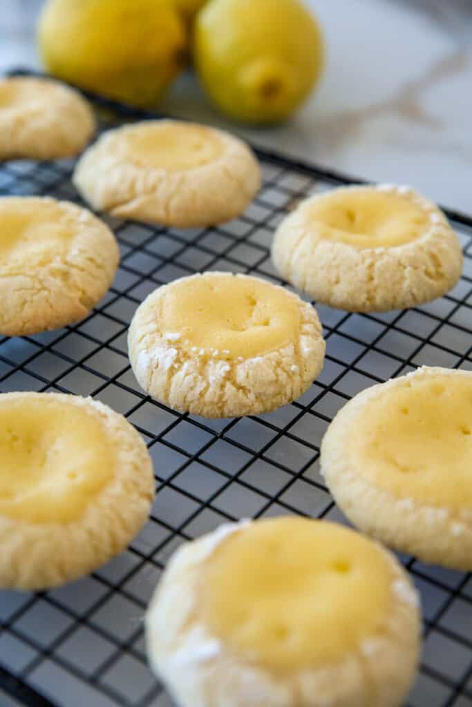 Cookies with a lemon filling are cooling on a black wire rack. In the background, there are two whole lemons slightly out of focus. The cookies have a crumbly texture with light powdered sugar dusting their edges.