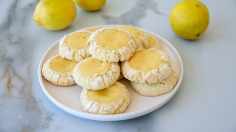 A plate of lemon crinkle cookies sits on a marble surface, surrounded by fresh lemons. The cookies are lightly dusted with powdered sugar, displaying a cracked texture and golden-yellow centers. The backdrop features more lemons, adding a bright, fresh ambiance.