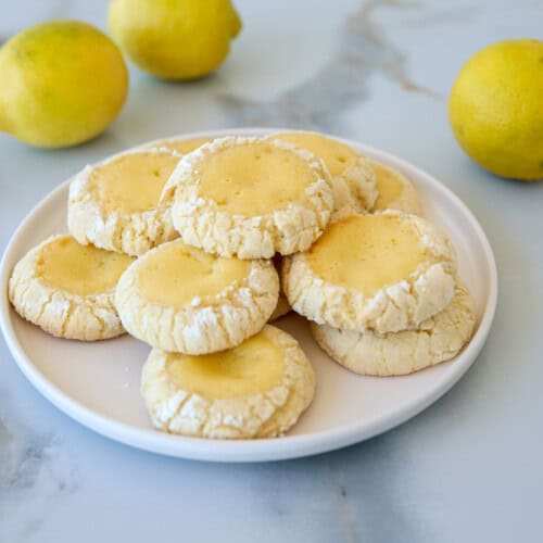 A plate of lemon crinkle cookies sits on a marble surface, surrounded by fresh lemons. The cookies are lightly dusted with powdered sugar, displaying a cracked texture and golden-yellow centers. The backdrop features more lemons, adding a bright, fresh ambiance.