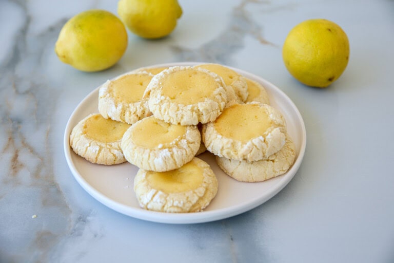 A plate of lemon crinkle cookies sits on a marble surface, surrounded by fresh lemons. The cookies are lightly dusted with powdered sugar, displaying a cracked texture and golden-yellow centers. The backdrop features more lemons, adding a bright, fresh ambiance.