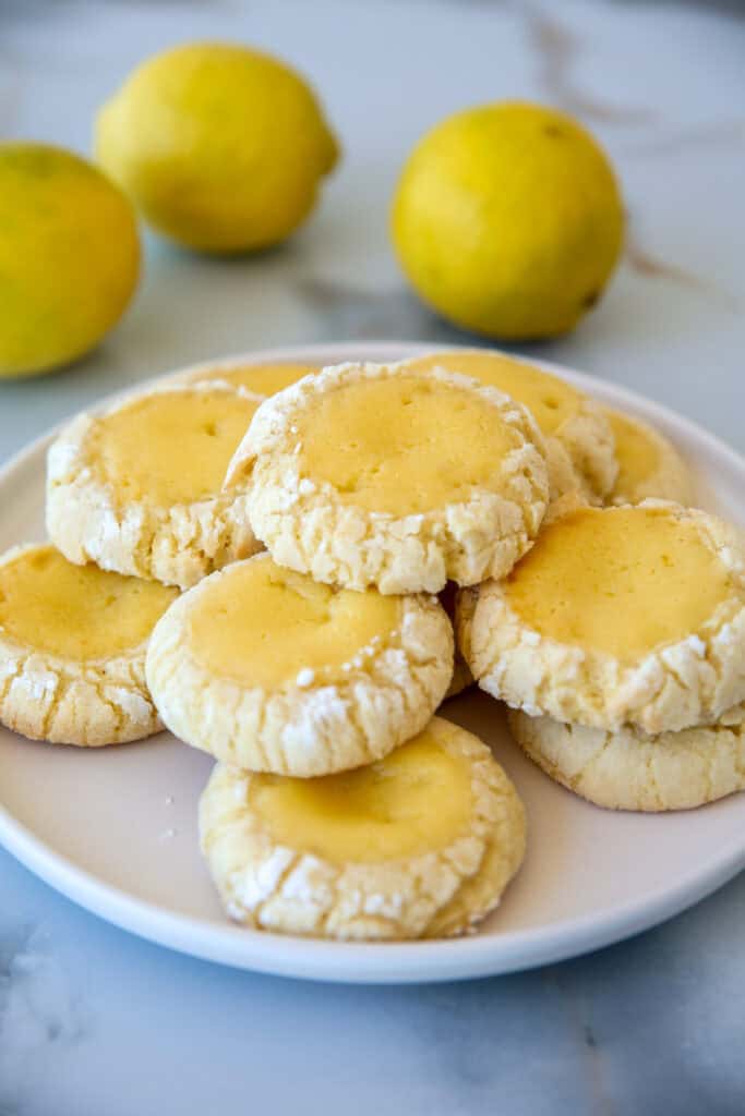 A plate of freshly baked lemon crinkle cookies is displayed, featuring a soft, yellow center with a slightly cracked, powdered sugar-dusted exterior. Three whole lemons are placed in the background on a marble surface.