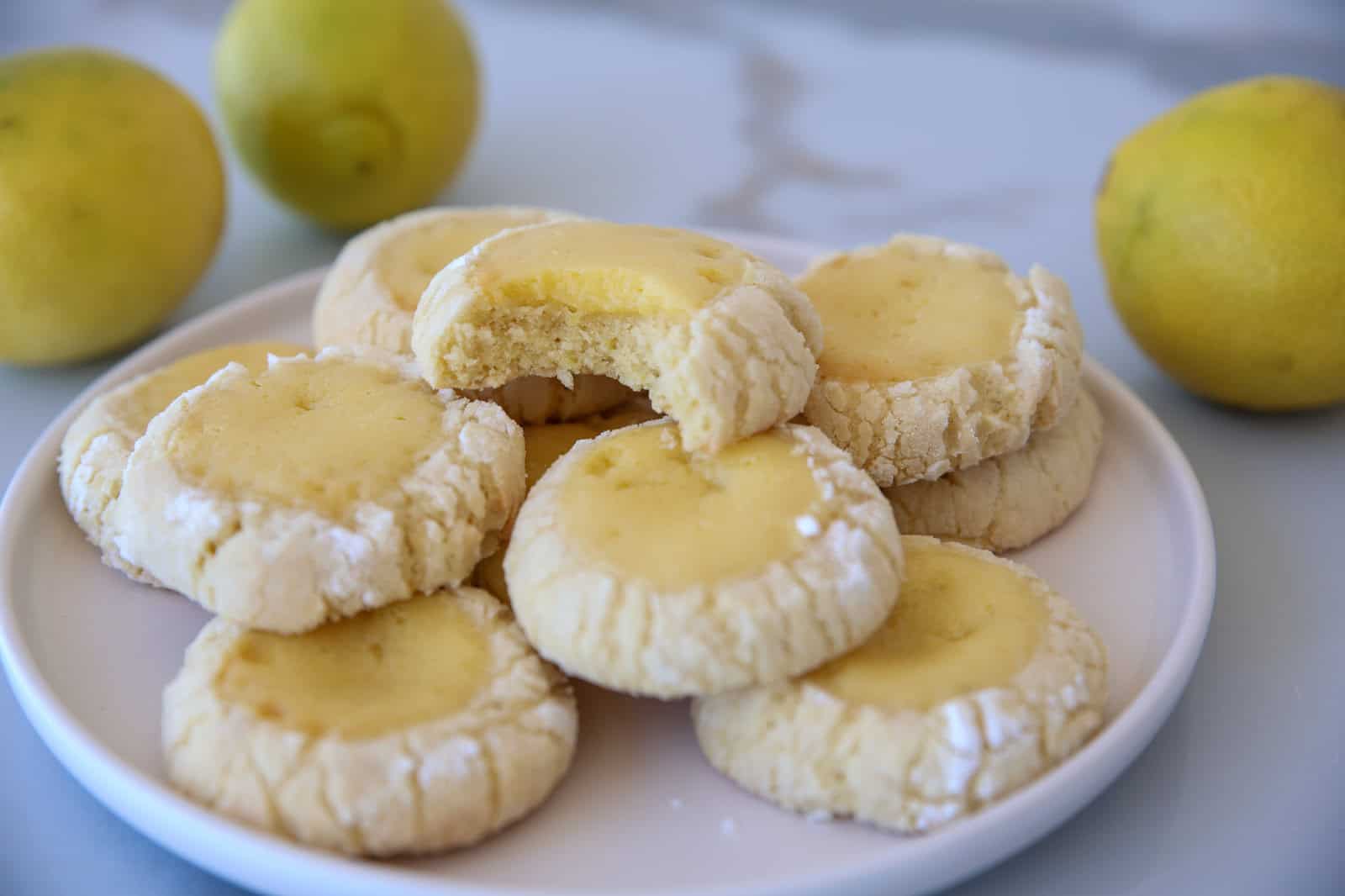 A white plate holds a stack of lemon crinkle cookies. The cookies have a powdered sugar coating and a cracked surface. One cookie on top has a bite taken out of it, revealing a soft, yellow interior. Fresh lemons are visible in the background on a light-colored table.