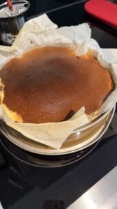 A freshly baked cheesecake with a dark, caramelized top sits in a round springform pan lined with parchment paper. The cake is placed on a black stovetop, and a red oven mitt is visible in the background.