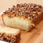 freshly baked maple cake with a golden brown crust and a topping of chopped nuts is placed on a wooden cutting board. A single slice has been cut from the loaf, revealing a soft and moist inside. The background is a wooden surface.