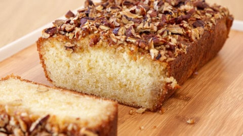 freshly baked maple cake with a golden brown crust and a topping of chopped nuts is placed on a wooden cutting board. A single slice has been cut from the loaf, revealing a soft and moist inside. The background is a wooden surface.