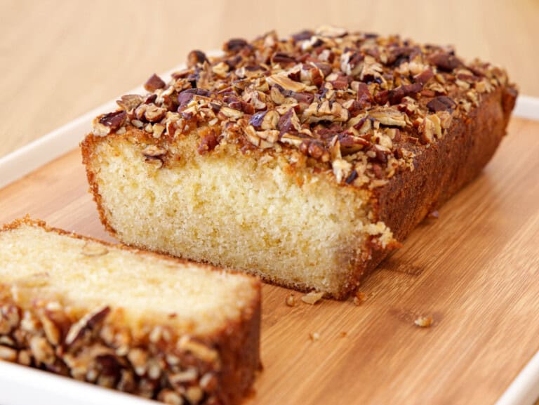 freshly baked maple cake with a golden brown crust and a topping of chopped nuts is placed on a wooden cutting board. A single slice has been cut from the loaf, revealing a soft and moist inside. The background is a wooden surface.