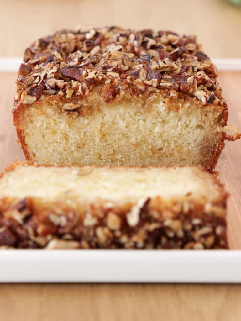 A close-up photo of a maple cake sliced to show its moist, light yellow interior. The cake is topped with a crunchy layer of chopped nuts. The loaf rests on a white rectangular plate, which sits on a wooden surface.