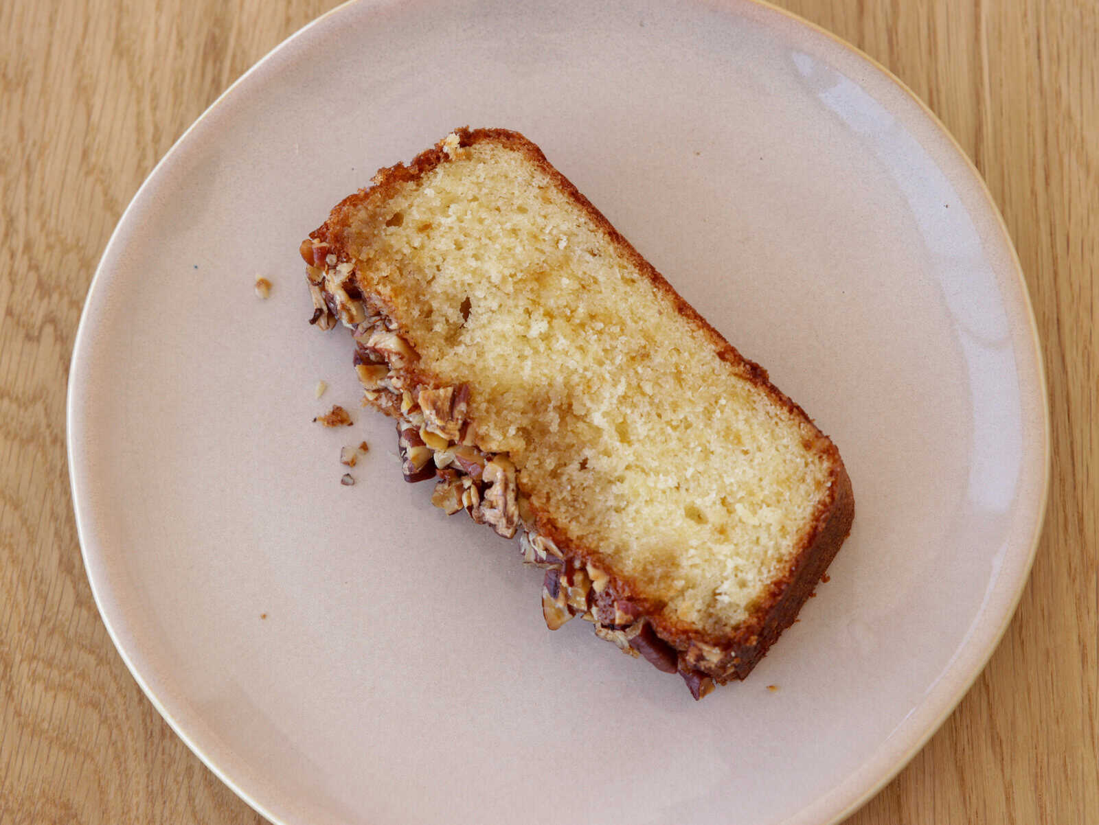 A slice of moist maple cake sits on a white plate placed on a wooden table. The cake has a golden-brown crust with a light yellow interior and is topped with a layer of chopped nuts, adding texture and flavor.