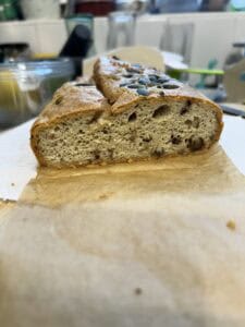A close-up of a sliced loaf of bread with seeds on top, sitting on brown parchment paper in a kitchen setting. The bread appears dense and grainy with visible air pockets and nuts inside.