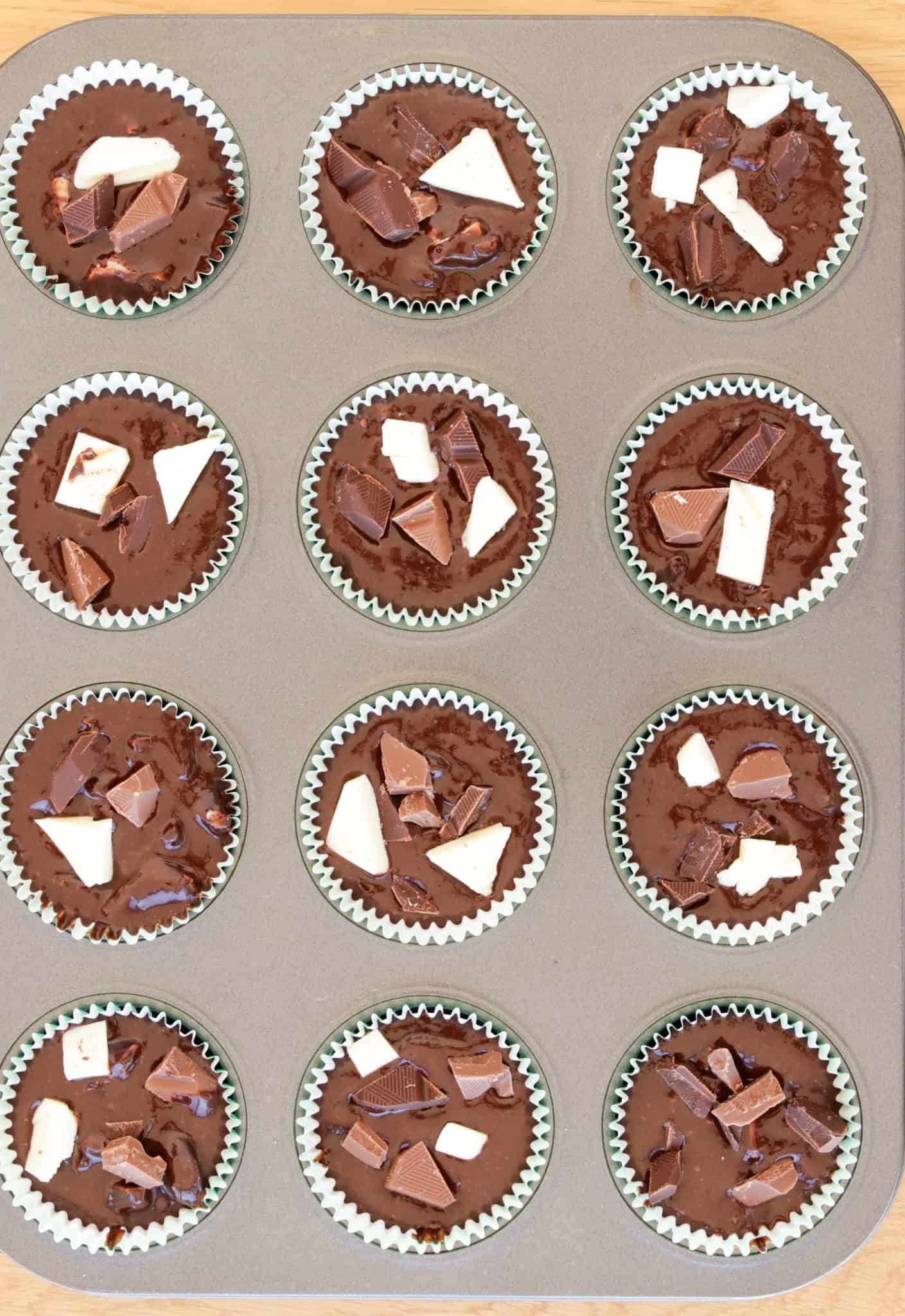 A muffin pan holds twelve chocolate muffins, each topped with various pieces of chopped chocolate and white chocolate. The muffins are in green paper liners and are about to be baked. The view is from above, showing the uniform arrangement.