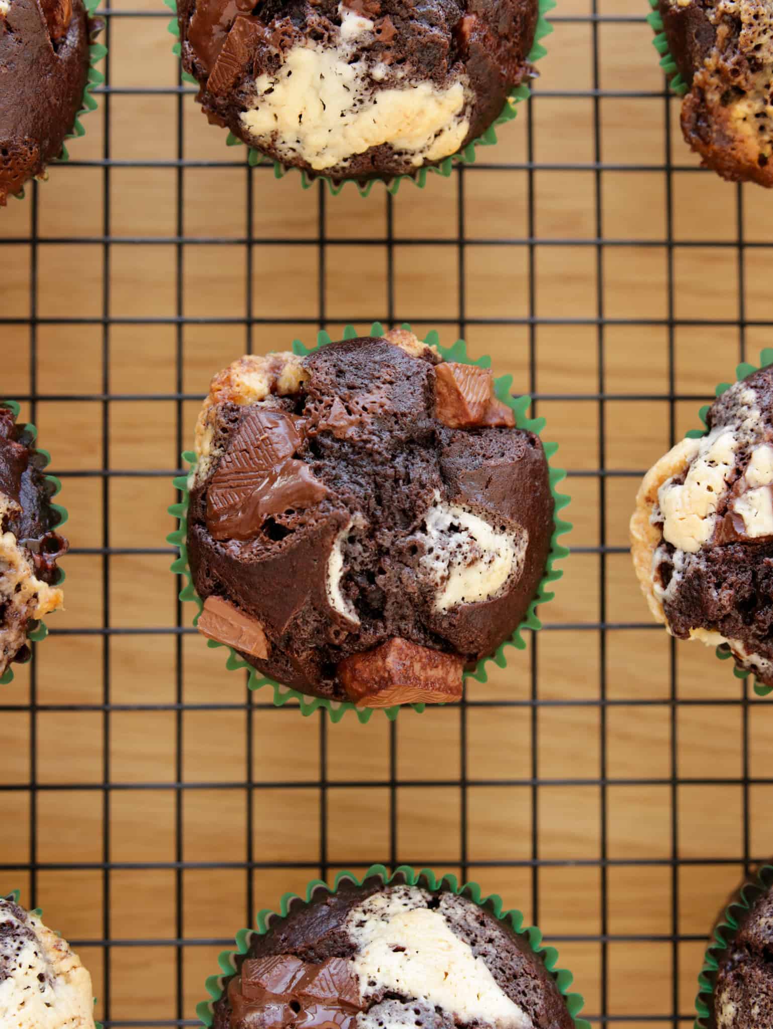 An overhead view of chocolate and vanilla marbled cupcakes with chunks of chocolate, displayed in green paper liners on a black wire cooling rack placed on a wooden surface.