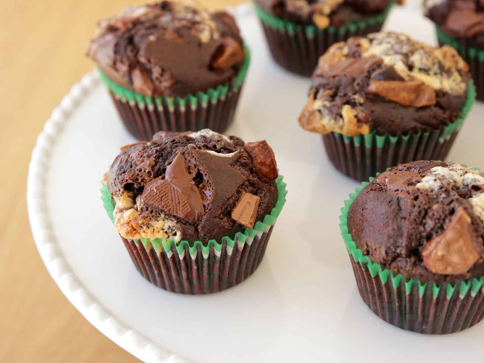 A close-up of several chocolate muffins with chunks of chocolate and possibly cream cheese swirls, wrapped in dark brown and green cupcake liners. They are arranged on a white plate with a scalloped edge, placed on a light wood surface.