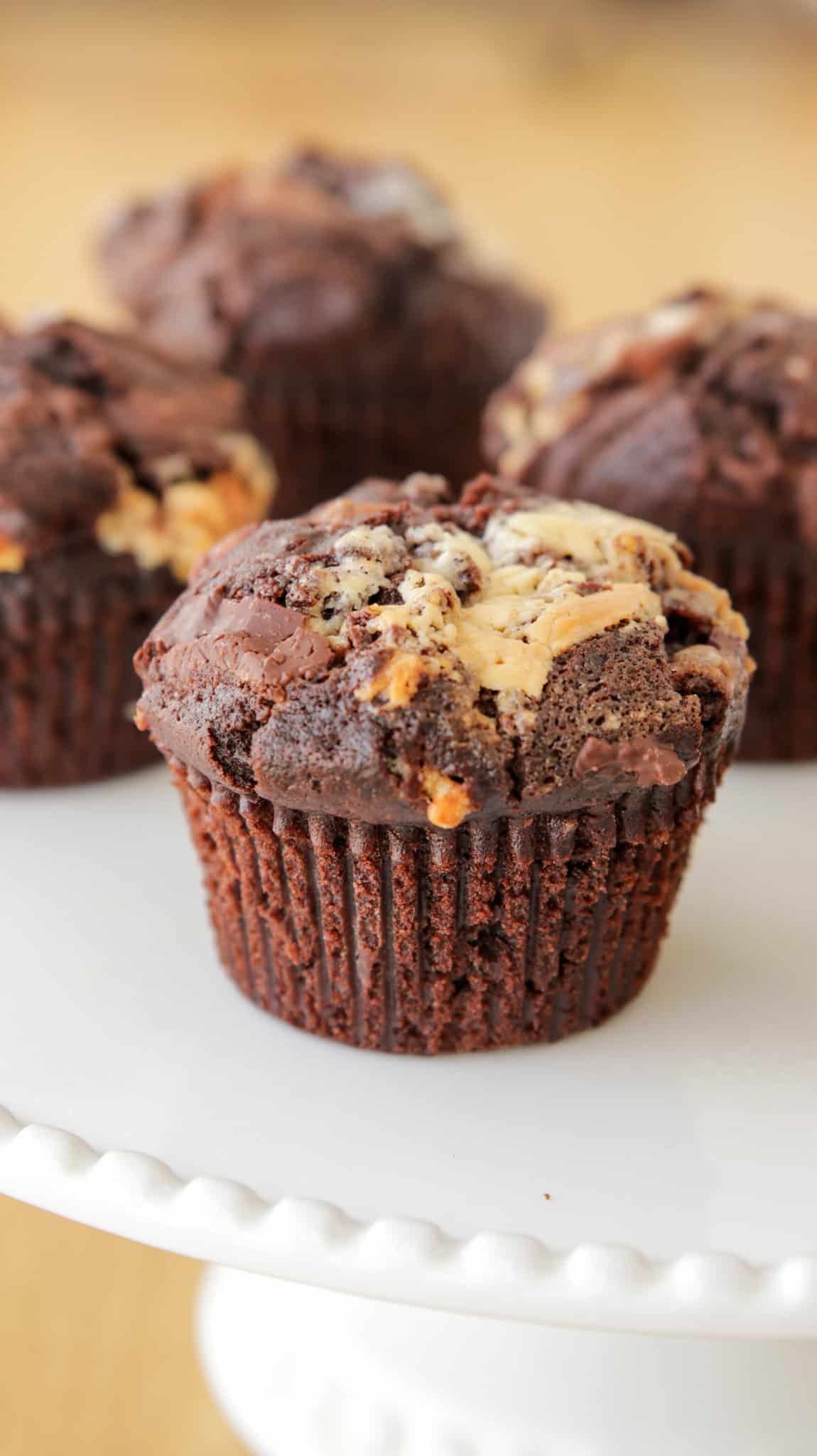 Close-up of a triple chocolate muffin with chunks of white chocolate on a white cake stand, with three additional muffins blurred in the background. The muffins appear moist and rich, with a slightly crumbled top.