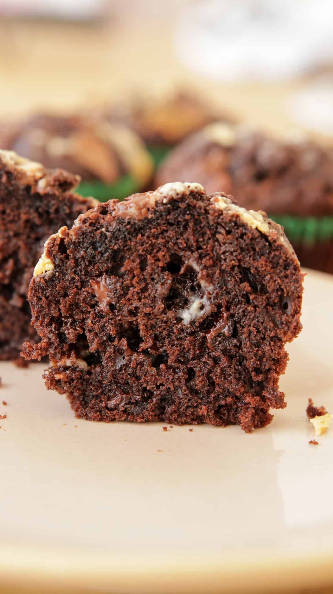 Close-up of a moist chocolate muffin cut in half, revealing its rich, dense texture and chocolate chunks throughout. The muffin is placed on a cream-colored plate, with more muffins blurred in the background.