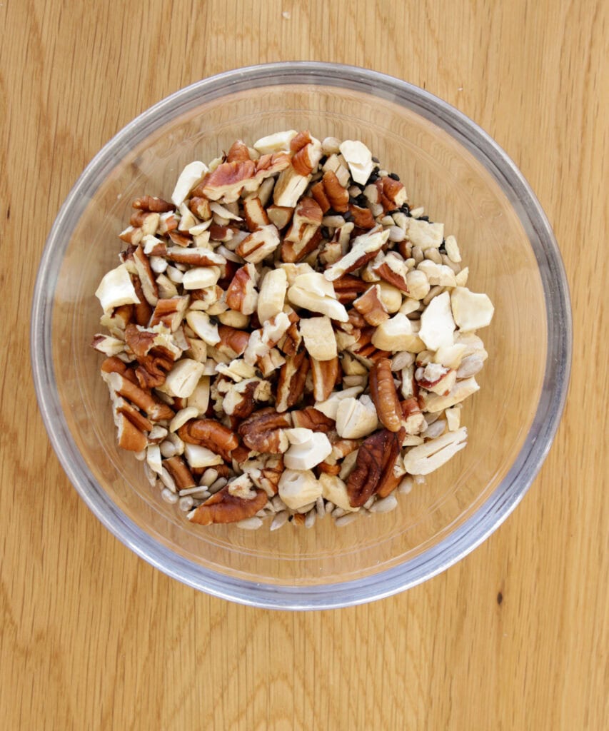 A clear glass bowl filled with a mix of chopped nuts, including pecans, cashews, and sunflower seeds, sits on a light wood surface.