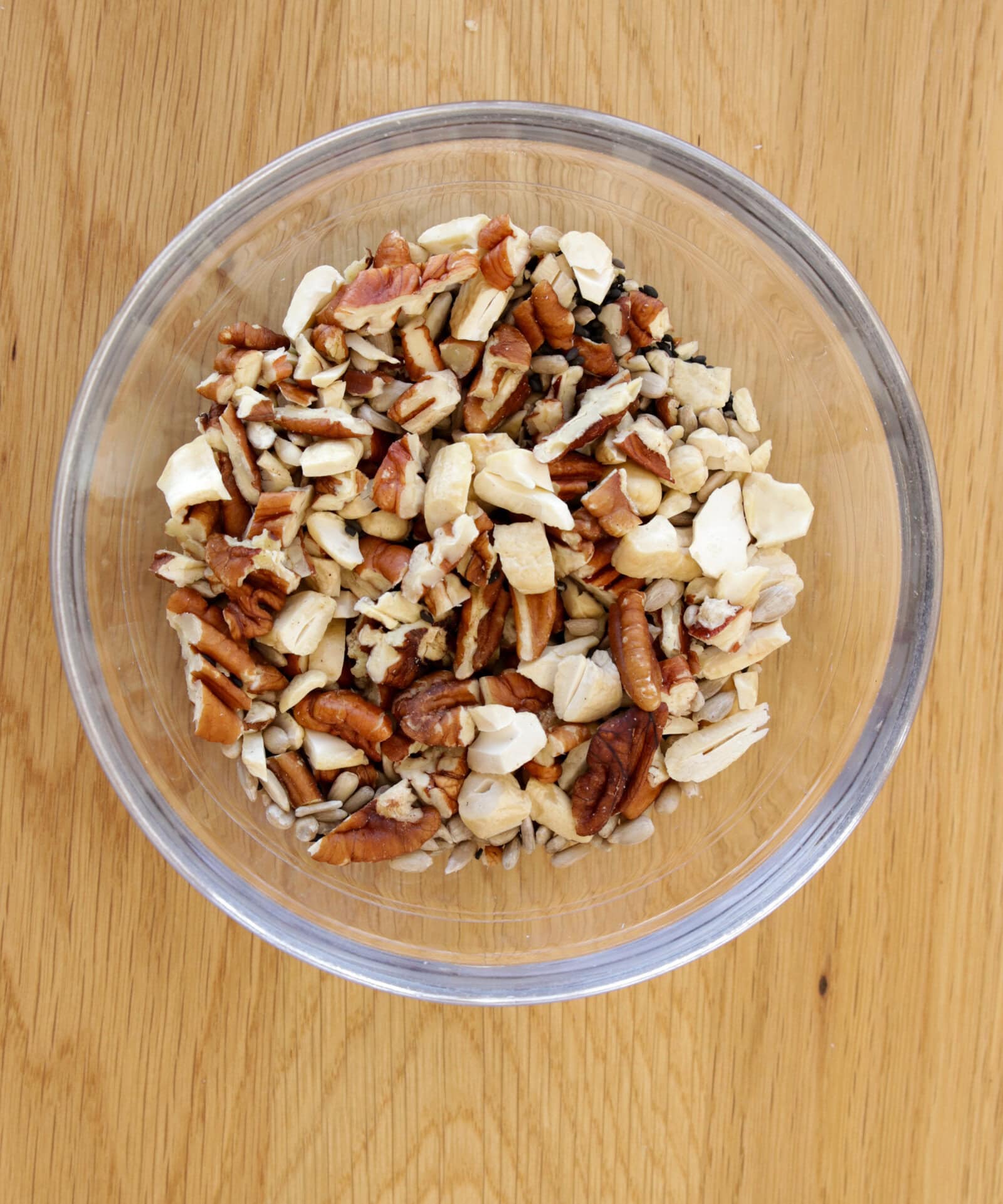 A clear glass bowl filled with a mix of chopped nuts, including pecans, cashews, and sunflower seeds, sits on a light wood surface.