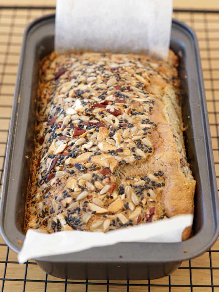 A freshly baked loaf of bread in a black rectangular baking tin, resting on a cooling rack. The bread is topped with a variety of seeds and nuts, and a piece of parchment paper is visible under one side of the loaf for easy removal.