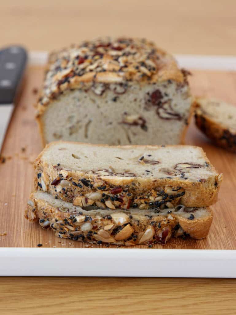 A sliced loaf of multigrain bread with visible seeds and nuts is placed on a white tray with a wooden cutting board. A bread knife rests beside the loaf. The background is a light-colored wooden surface.