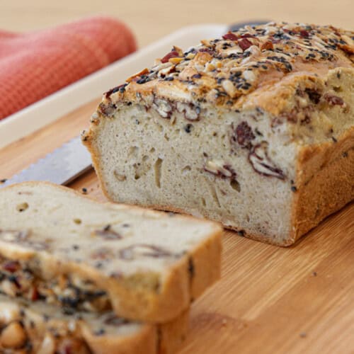 A flourless seeded loaf of bread sits on a wooden cutting board with slices cut from the end, revealing a fluffy interior with visible nuts and seeds. A serrated knife and a red cloth are placed beside the bread.