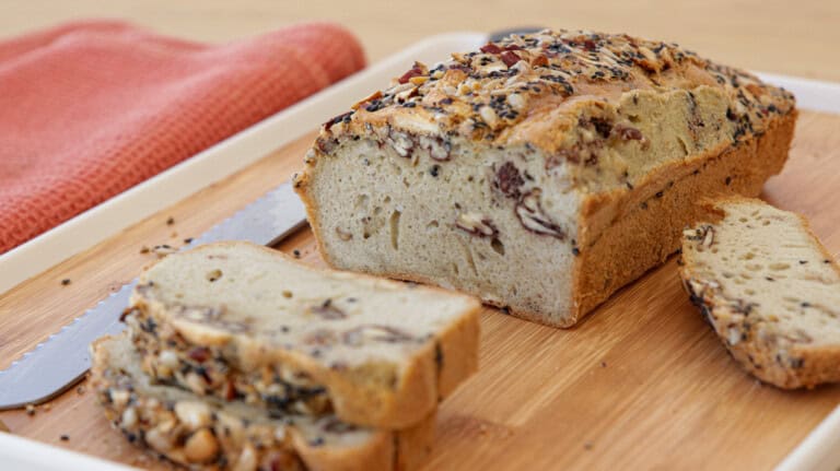 A flourless seeded loaf of bread sits on a wooden cutting board with slices cut from the end, revealing a fluffy interior with visible nuts and seeds. A serrated knife and a red cloth are placed beside the bread.