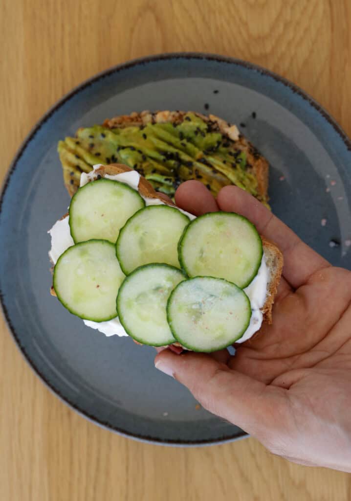 A person holding a slice of bread topped with cucumber slices and white spread over a gray plate with another slice of bread topped with mashed avocado and seeds. The background is a wooden surface.