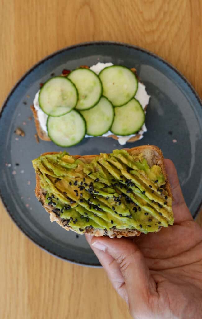 A hand holding a slice of toast topped with mashed avocado and black sesame seeds. In the background, a gray plate holds another slice of toast topped with cream cheese and cucumber slices. The scene is set on a light wooden surface.