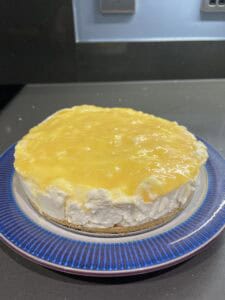A homemade cheesecake with a biscuit base, topped with a glossy yellow lemon curd layer, sits on a blue-rimmed plate on a dark kitchen countertop.