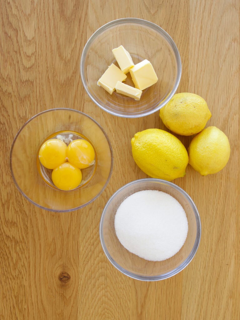 Four bowls of ingredients on a wooden surface: three egg yolks in one bowl, butter cubes in another, three whole lemons beside them, and a bowl of granulated sugar.