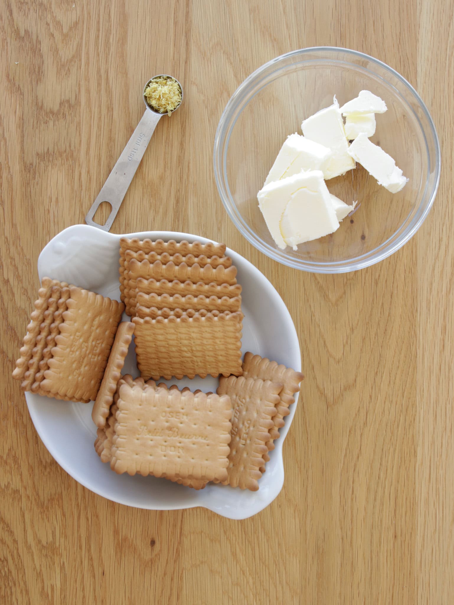 An overhead view of a wooden surface with a white dish containing rectangular biscuits, a metal measuring spoon filled with a small amount of yellowish substance, and a clear bowl holding pieces of butter.