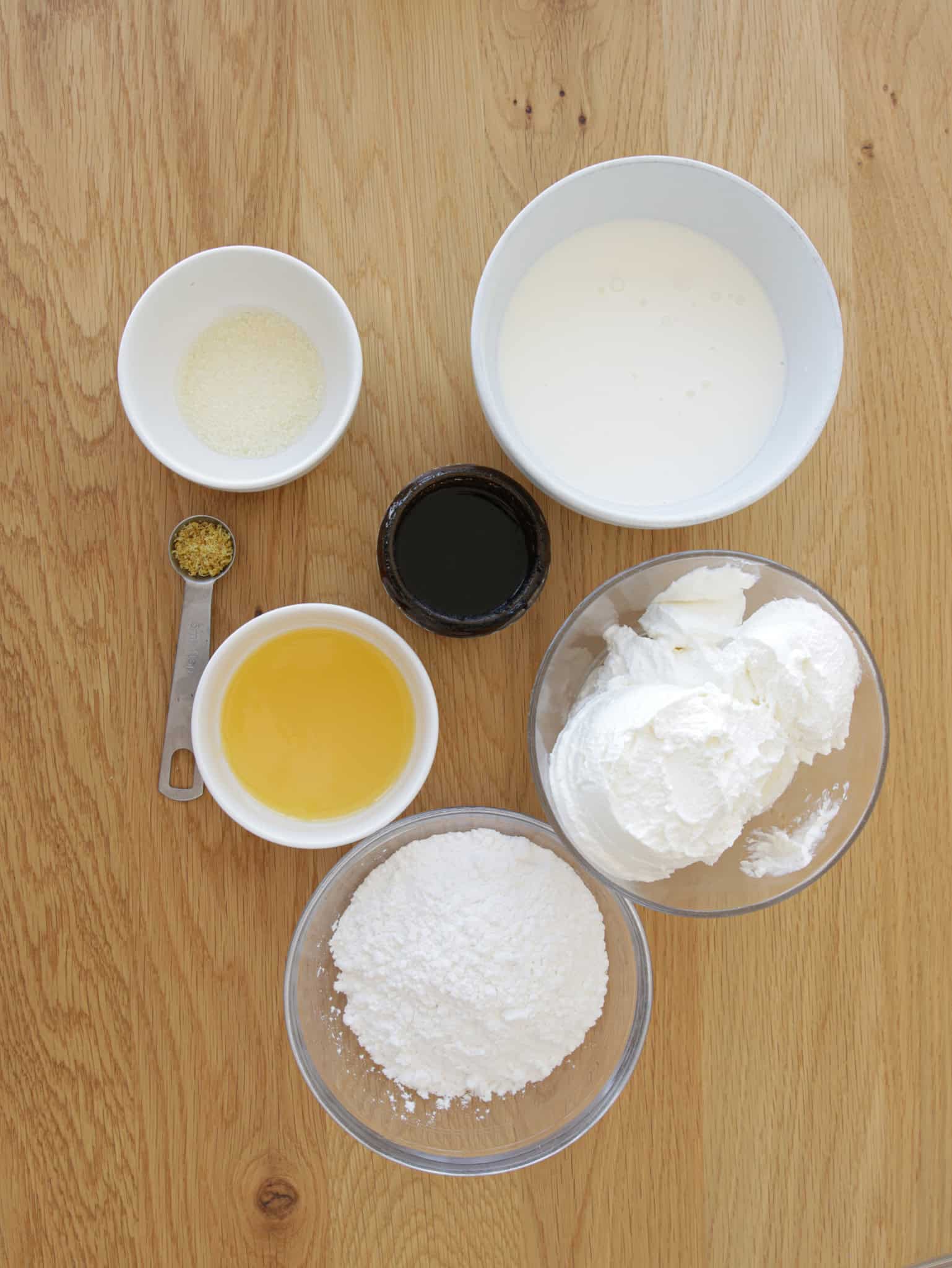 Top-down view of six bowls and a measuring spoon with various ingredients on a wooden surface. Visible items include powdered sugar, ricotta cheese, lemon zest, honey, vanilla extract, milk, and granulated sugar.