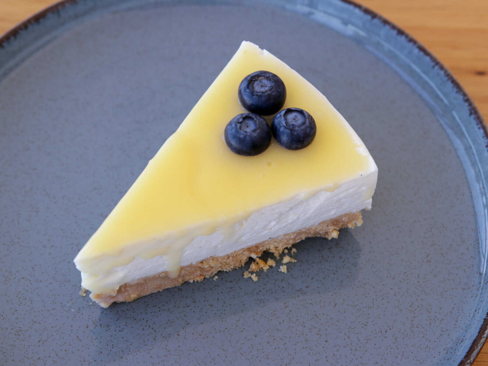 A slice of lemon cheesecake on a gray plate, topped with three fresh blueberries. The cheesecake features a creamy lemon layer, a white creamy middle layer, and a crumbly crust at the bottom.