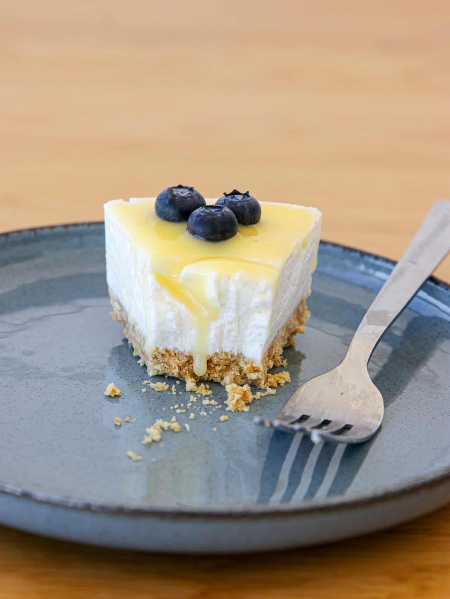 A slice of cheesecake with a graham cracker crust sits on a blue plate. The cheesecake is topped with yellow lemon glaze and three blueberries. A stainless steel fork lies beside the slice on the plate. Crumbs are scattered near the fork.