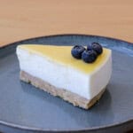 A slice of cheesecake with a smooth, yellow topping is garnished with three blueberries. The cheesecake rests on a crumbly biscuit base and is served on a simple, round, grayish-blue plate on a light wooden surface.