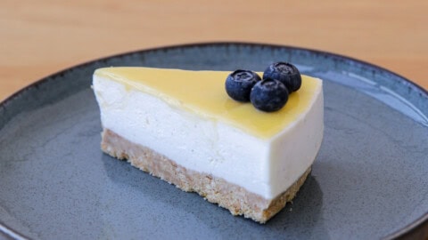 A slice of cheesecake with a yellow topping, garnished with three blueberries on top, is served on a round, gray plate. The cake has a crumbly base, a creamy middle layer, and a smooth, glossy top layer. The background is a light wooden surface.