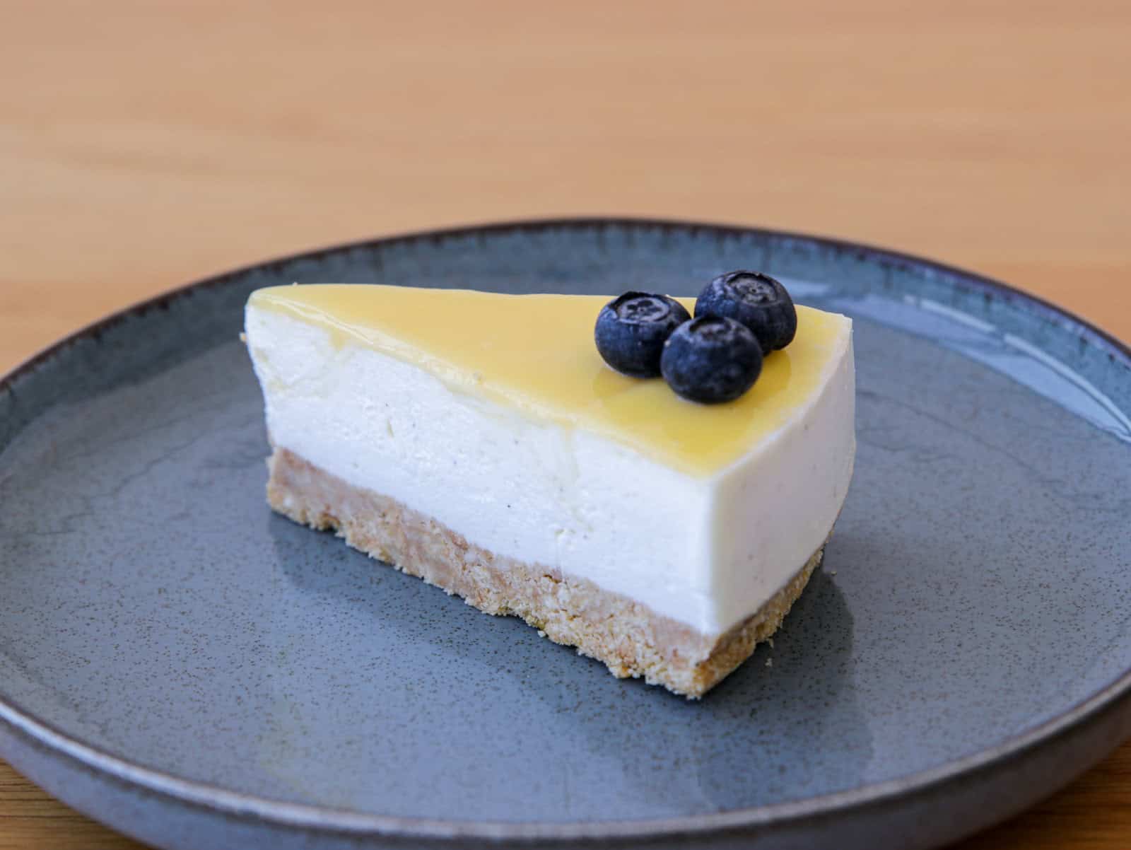 A slice of cheesecake with a yellow topping, garnished with three blueberries on top, is served on a round, gray plate. The cake has a crumbly base, a creamy middle layer, and a smooth, glossy top layer. The background is a light wooden surface.