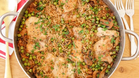 A large skillet filled with a colorful dish of cooked chicken thighs, rice, peas, mushrooms, and herbs. The meal is garnished with chopped parsley, adding a touch of green. Two forks and a striped cloth are visible next to the skillet on a wooden table.