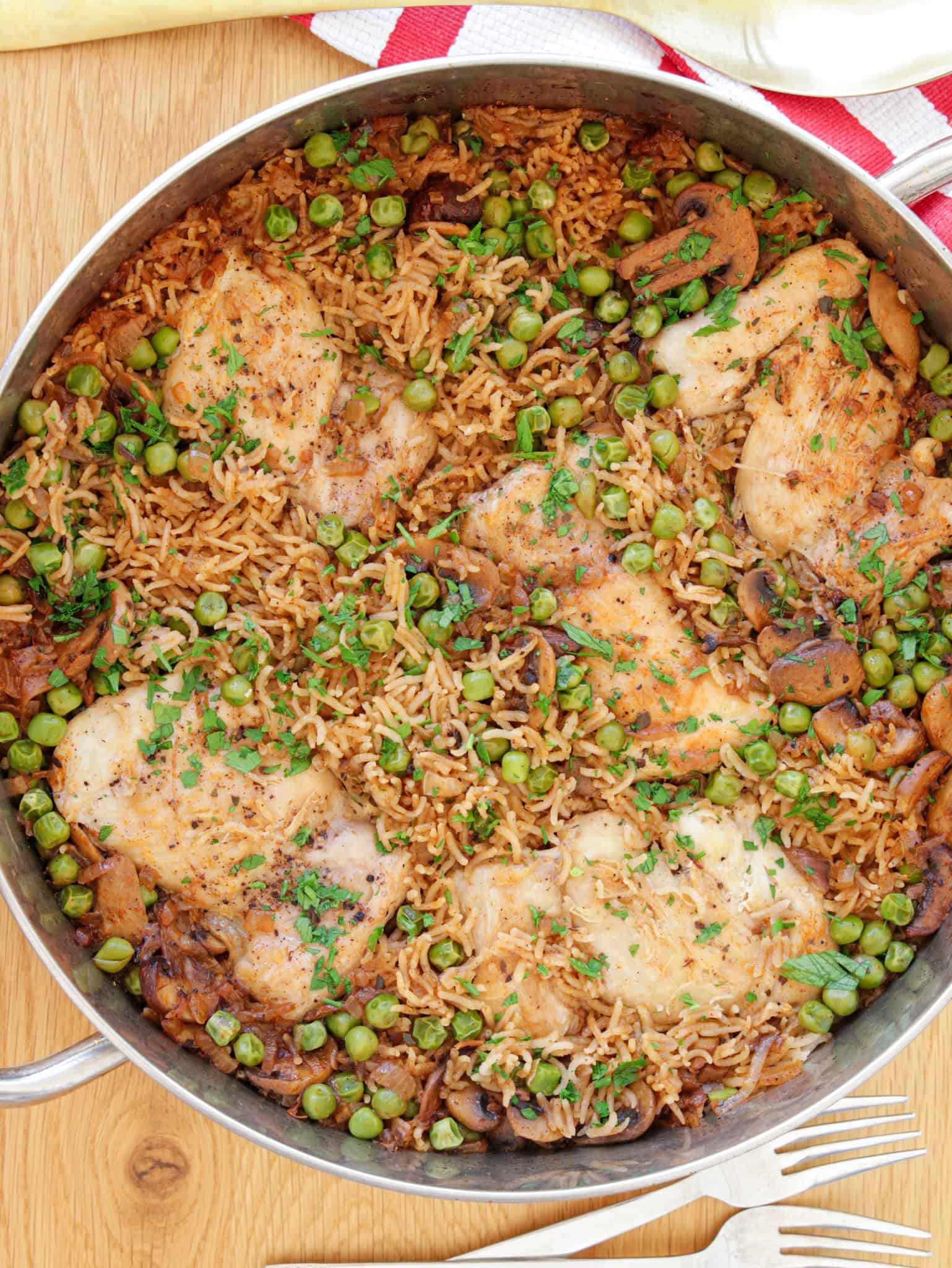 A skillet filled with a chicken and rice dish, including pieces of chicken thighs, peas, mushrooms, and rice seasoned with herbs. The meal is presented on a wooden surface with a red and white cloth partially visible at the top, accompanied by silver utensils.