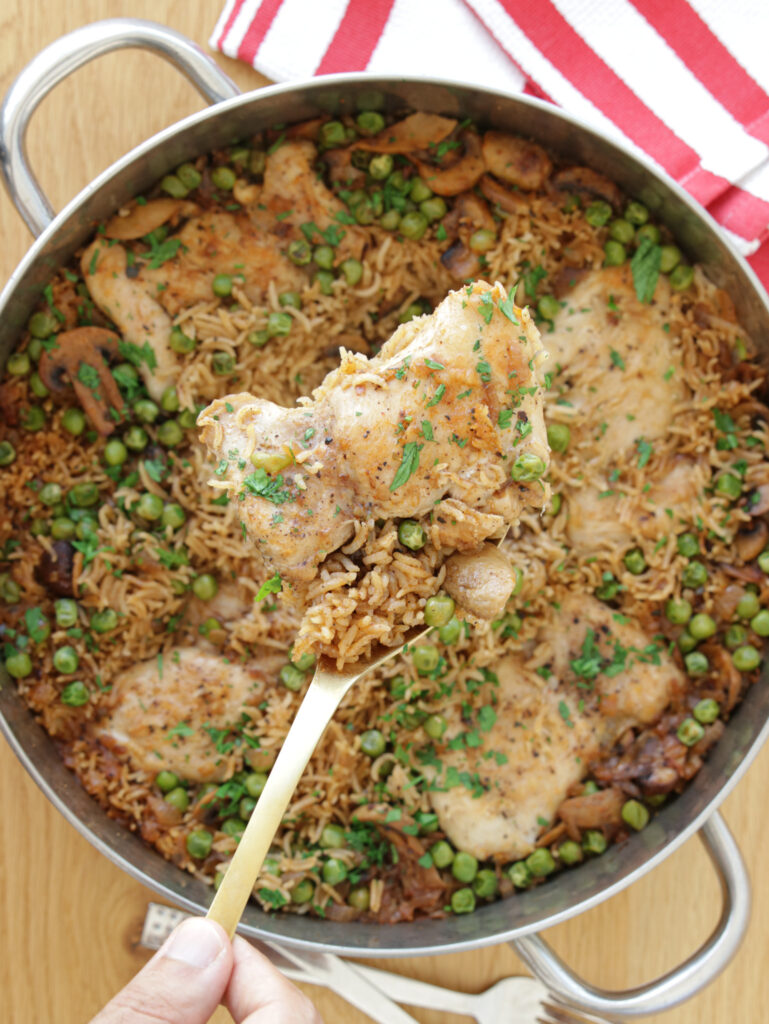 A round pan filled with a cooked one-pot meal consisting of seasoned chicken thighs, rice, peas, and mushrooms. A hand holds a golden fork, lifting a piece of chicken and rice from the pan. A striped red and white cloth is partially visible in the background.