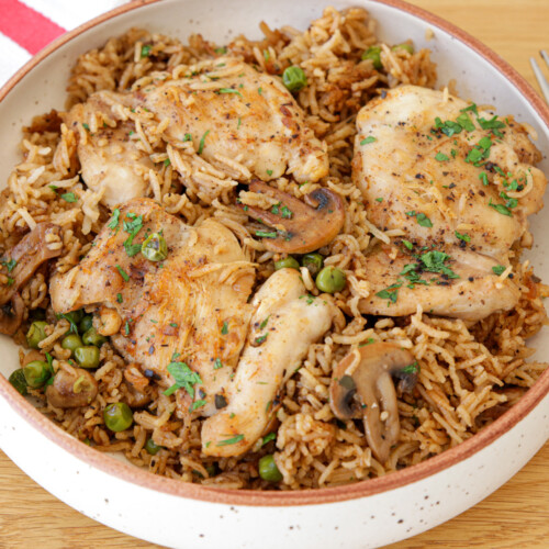 A bowl of seasoned rice topped with cooked chicken thighs, mushrooms, peas, and garnished with fresh herbs. A red and white striped cloth and a fork are placed beside the bowl on a wooden surface.