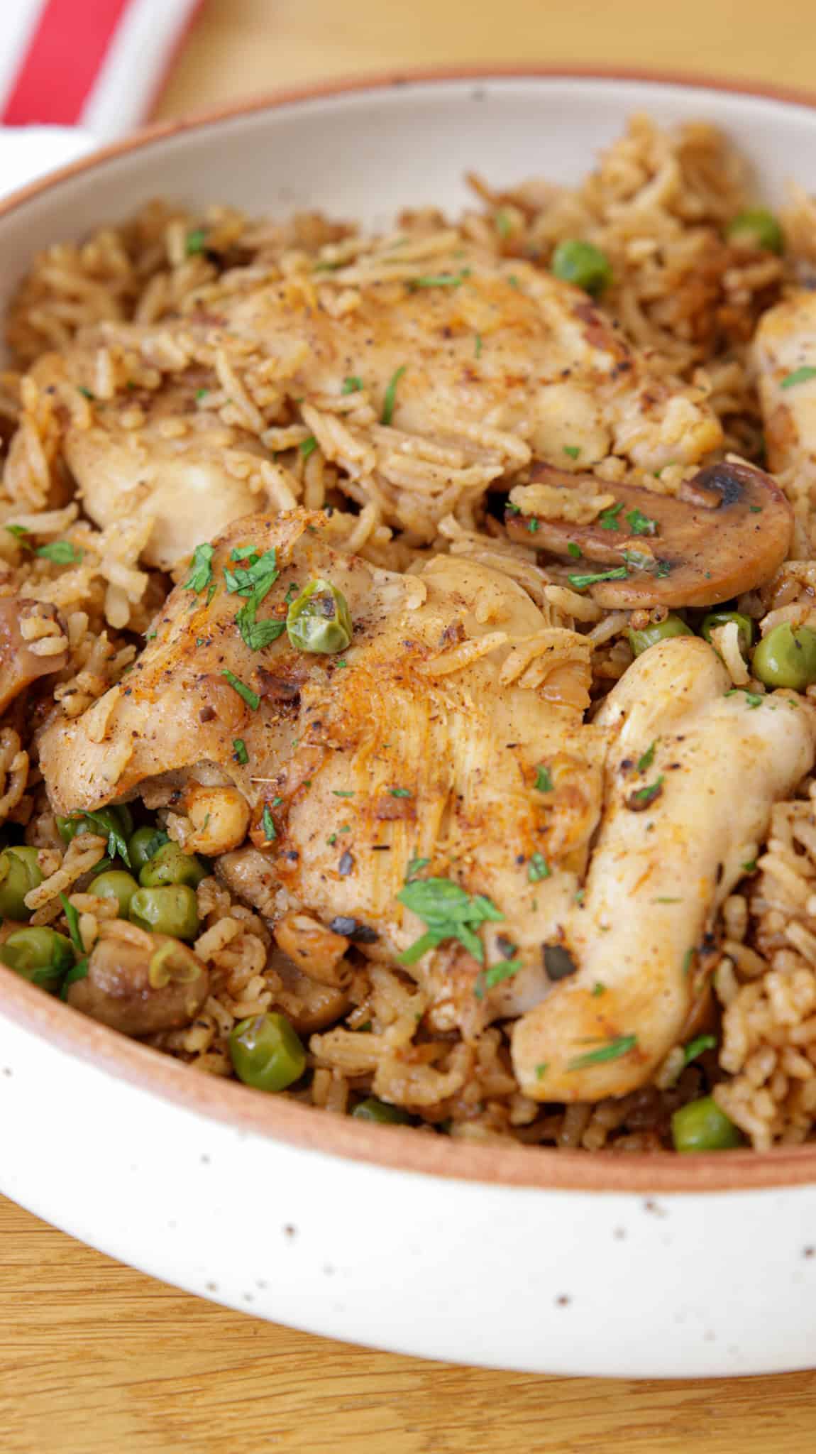 A close-up of a bowl filled with a savory dish consisting of pieces of grilled chicken atop a bed of rice mixed with green peas and mushrooms. The dish is garnished with chopped herbs and has a slightly golden, appetizing appearance.