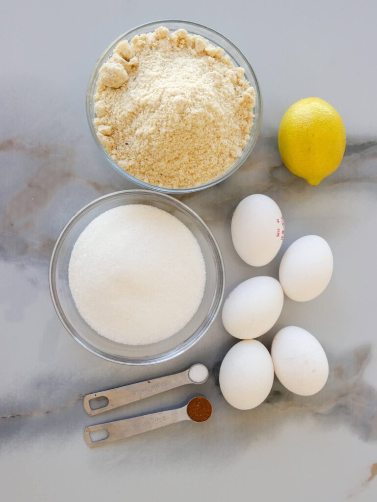 ingredients for tarta de Santiago: including a bowl of almond flour, a bowl of granulated sugar, four eggs, a whole lemon, a teaspoon of baking powder, and a teaspoon of coffee grounds on a marble surface.