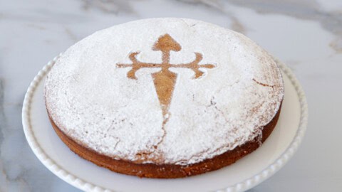 Tarta de Santiago A round almond cake, dusted with powdered sugar, sits on a white plate. The cake has the emblem of the Cross of Saint James stenciled in the center using a contrasting caramel-colored powdered sugar design. The background is a marble surface.