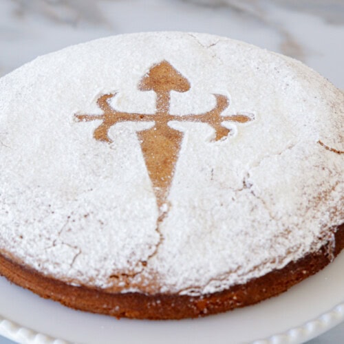 Tarta de Santiago A round almond cake, dusted with powdered sugar, sits on a white plate. The cake has the emblem of the Cross of Saint James stenciled in the center using a contrasting caramel-colored powdered sugar design. The background is a marble surface.