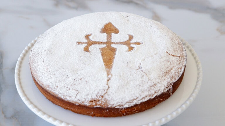 Tarta de Santiago A round almond cake, dusted with powdered sugar, sits on a white plate. The cake has the emblem of the Cross of Saint James stenciled in the center using a contrasting caramel-colored powdered sugar design. The background is a marble surface.