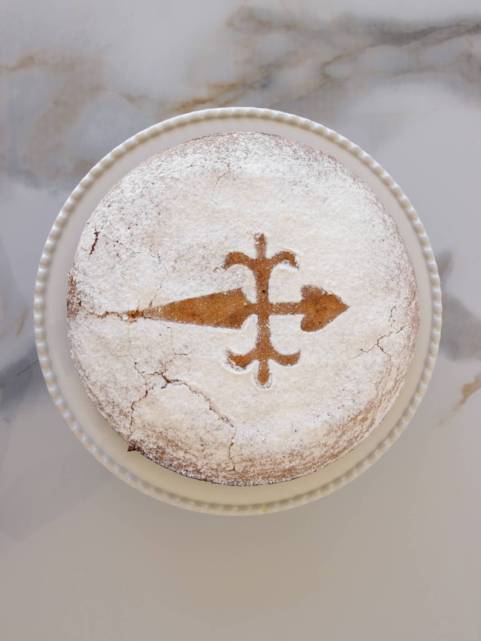 Spanish almond cake: A round cake dusted with powdered sugar, featuring a cross design prominently in the center. The cake is placed on a white ceramic plate with a beaded edge, on a marbled surface. The cake has visible cracks on the surface.