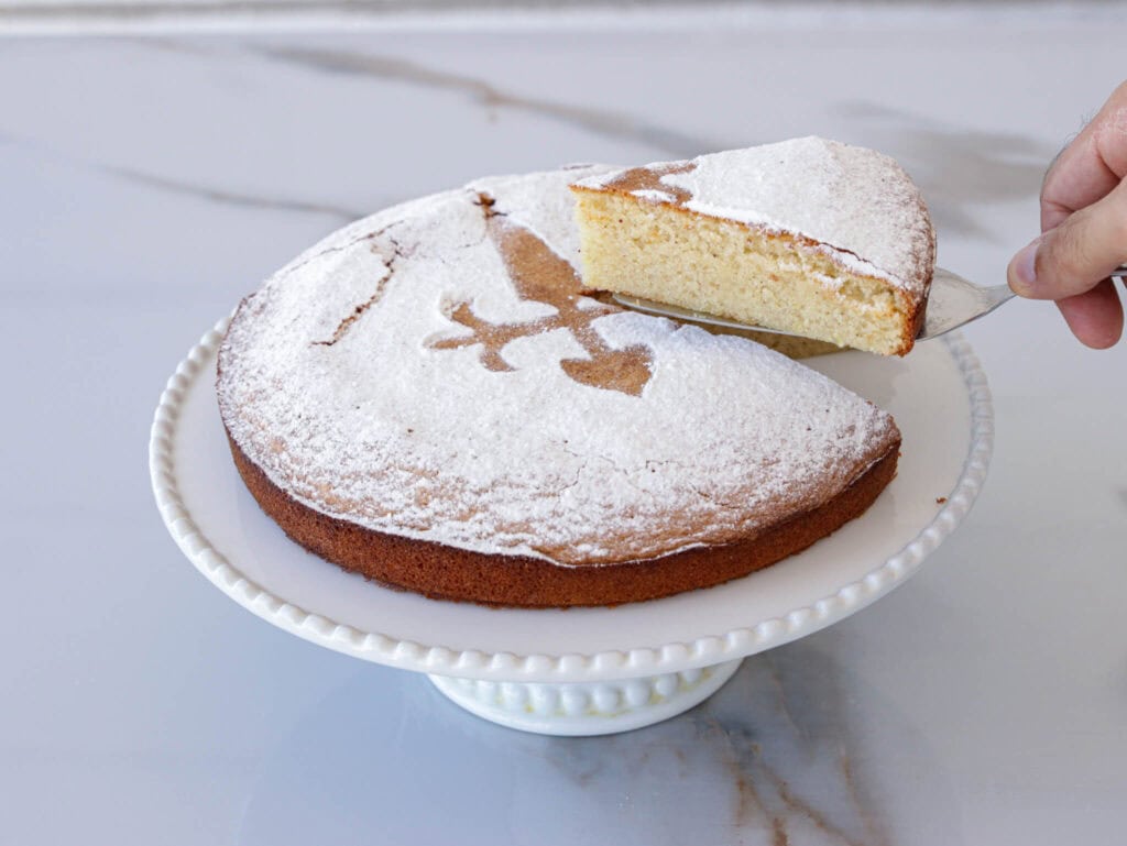 tarta de Santiago: A hand is lifting a slice of a round cake dusted with powdered sugar from a white cake stand. The cake has a decorative pattern on top made by the absence of powdered sugar, revealing the plain cake surface underneath. The background is a marble countertop.