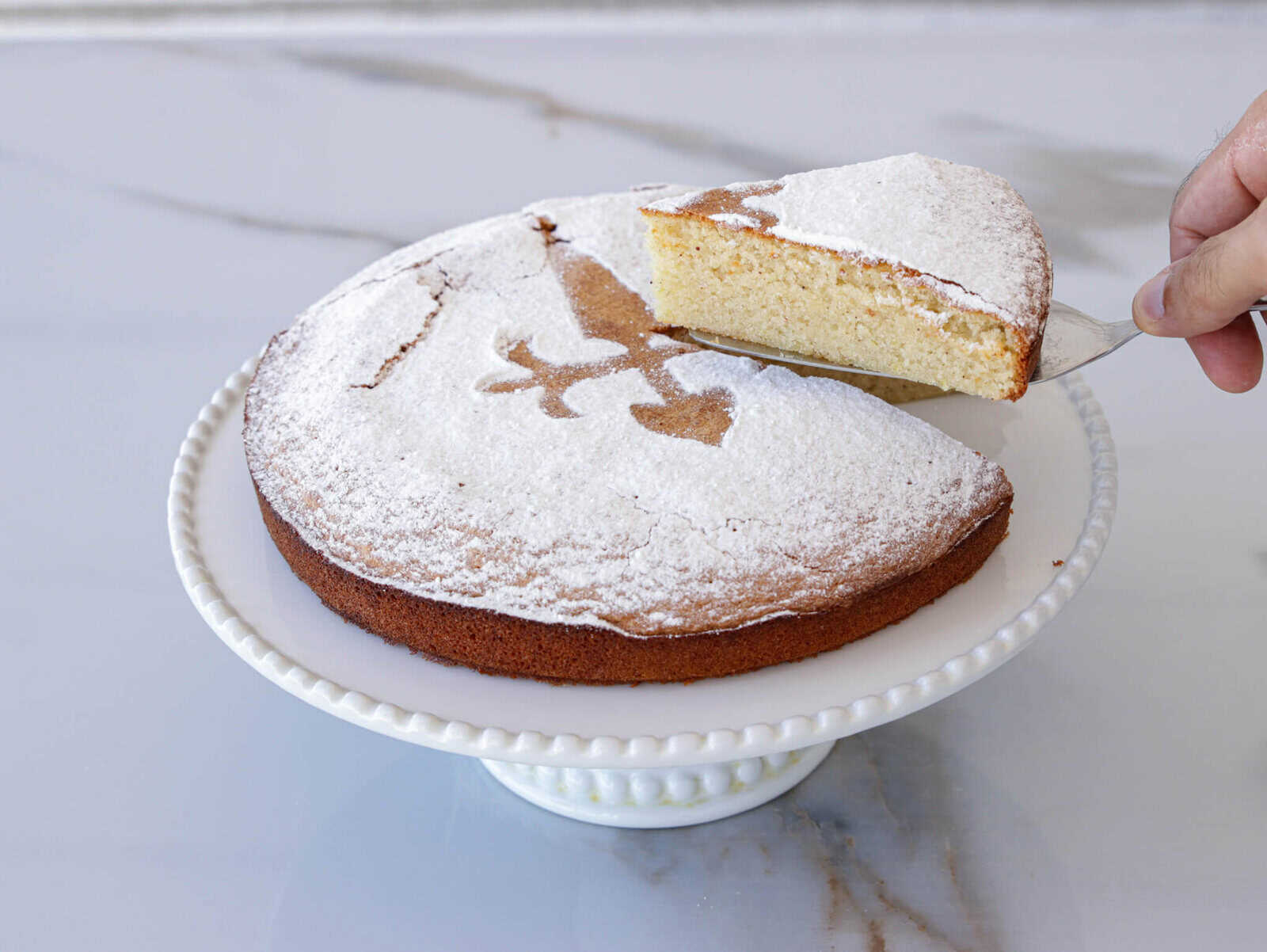 tarta de Santiago: A hand is lifting a slice of a round cake dusted with powdered sugar from a white cake stand. The cake has a decorative pattern on top made by the absence of powdered sugar, revealing the plain cake surface underneath. The background is a marble countertop.