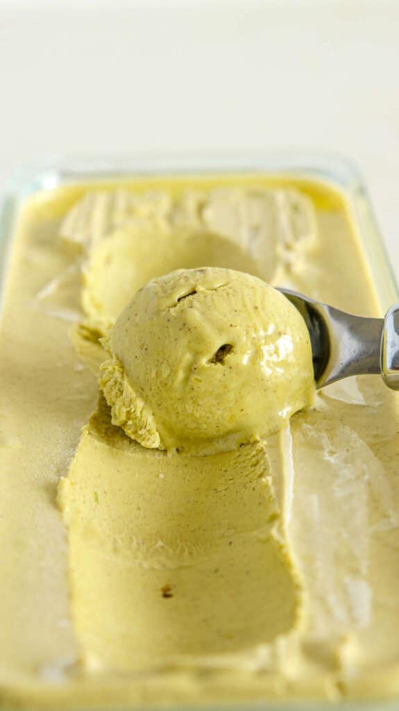 A metal ice cream scoop is lifting a round scoop of creamy, pale yellow ice cream from a rectangular glass container filled with the same ice cream. The ice cream has a smooth texture and appears freshly made.